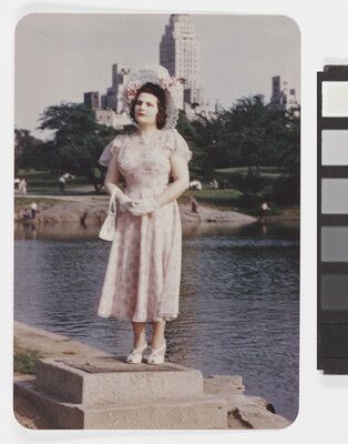 A color photograph of a White woman wearing a decorative sunhat and a floral dress standing on a concrete block beside a pond with tall buildings in the background.