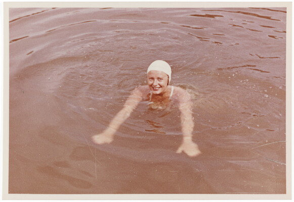 A color photograph of a smiling White woman wearing a white swim cap and swimming in what looks like a lake.