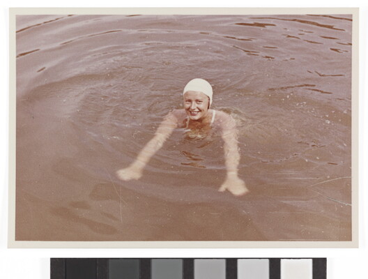 A color photograph of a smiling White woman wearing a white swim cap and swimming in what looks like a lake.