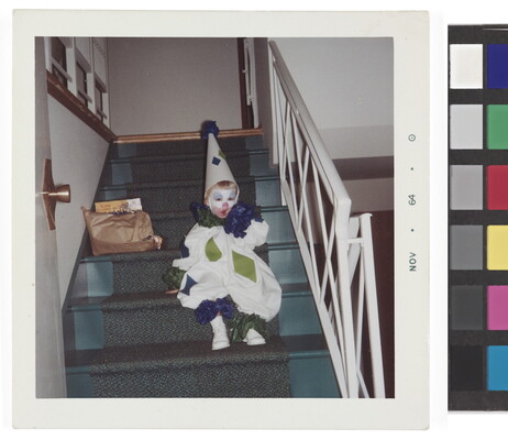 A color snapshot of a toddler in a blue, green, and white clown costume sitting on stairs inside a house.