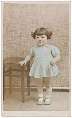 A color studio photograph of a White toddler with dark bobbed hair wearing a light blue dress and shoes leaning against a small table.