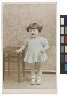 A color studio photograph of a White toddler with dark bobbed hair wearing a light blue dress and shoes leaning against a small table.