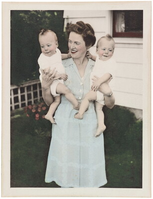A color photograph of a White woman wearing a light blue dress holding two babies, one in each arm.