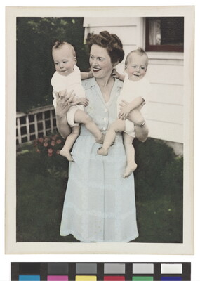 A color photograph of a White woman wearing a light blue dress holding two babies, one in each arm.