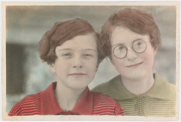A color photograph of two White children with short auburn hair, the child on the right wears glasses, leaning their heads toward each other.