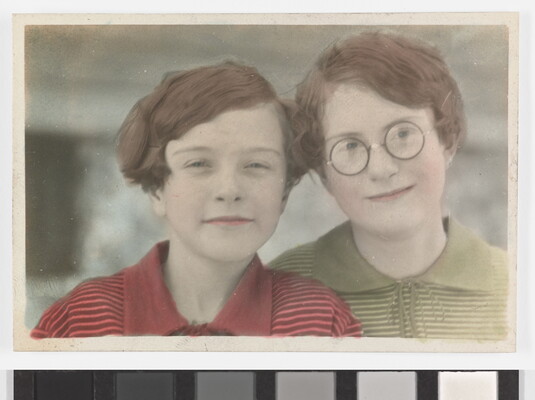 A color photograph of two White children with short auburn hair, the child on the right wears glasses, leaning their heads toward each other.