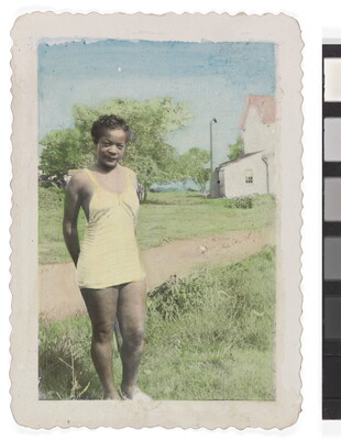 A color photograph of a young Black woman wearing a yellow one-piece bathing suit standing in grass beside a dirt road.