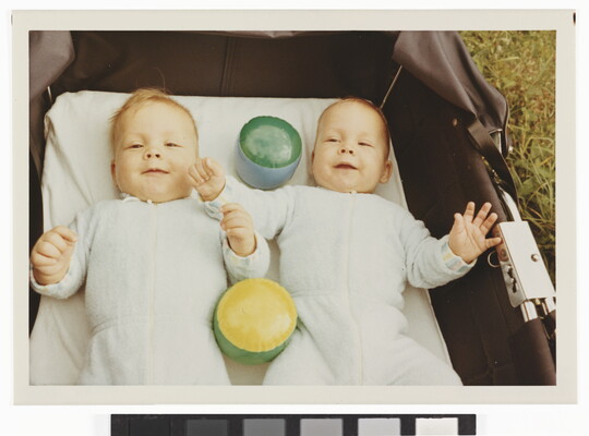 A color photograph of two White babies smiling and lying on their backs with toys in a baby carriage that is sitting in grass.