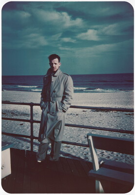 A color photograph of a White man in a long trench coat leaning against a railing with a beach in the background.