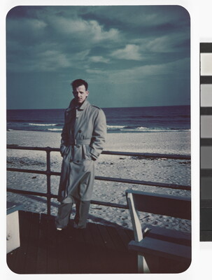 A color photograph of a White man in a long trench coat leaning against a railing with a beach in the background.