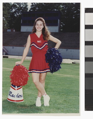 A color photograph of a young, smiling White woman in a red cheerleader uniform, holding pom-poms and leaning on a megaphone on a grassy sports field.
