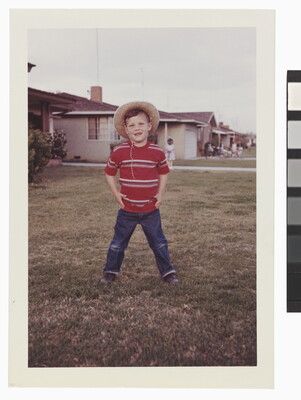 A color snapshot photograph of a young White child standing in a yard wearing a cowboy hat, red striped shirt, and blue jeans.
