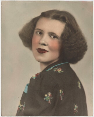 A color portrait photograph of a White woman with curly bobbed hair, wearing a floral top looking over her shoulder at the camera.
