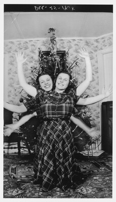 A black-and-white photograph of two young White women, both wearing plaid dresses, standing one behind the other in front of a Christmas tree waving their arms so it looks like they have two heads and six arms.