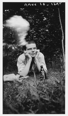 A black-and-white photograph of young White man in glasses, lying on his stomach in grass, his head propped in his hands; a beer bottle is in front of him, and a cloud of cigarette smoke hovers above his head.