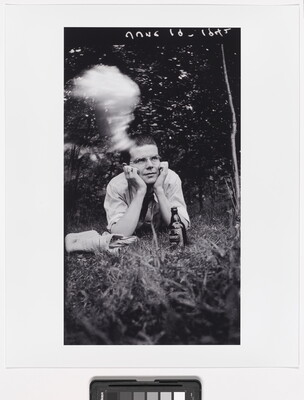 A black-and-white photograph of young White man in glasses, lying on his stomach in grass, his head propped in his hands; a beer bottle is in front of him, and a cloud of cigarette smoke hovers above his head.