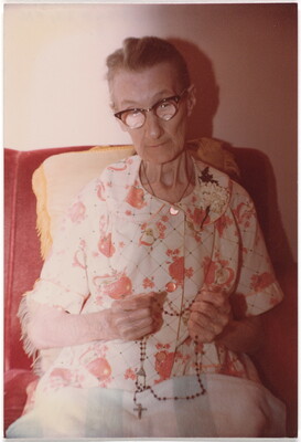 A color photograph of a seated elderly White woman, gray hair pulled back, wearing horn-rimmed glasses and a floral dress, a rosary in her hands.