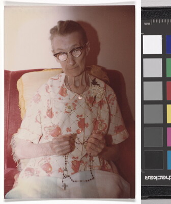 A color photograph of a seated elderly White woman, gray hair pulled back, wearing horn-rimmed glasses and a floral dress, a rosary in her hands.