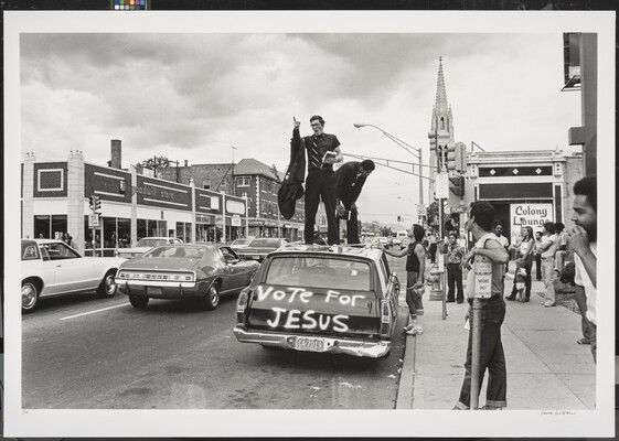 A black-and-white photograph of a downtown street scene of a man preaching from the top of a car with "Vote for Jesus" spray-painted on the back.