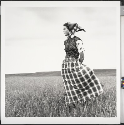 A black-and-white photograph of a young White woman wearing a long plaid skirt and a headscarf standing in field.