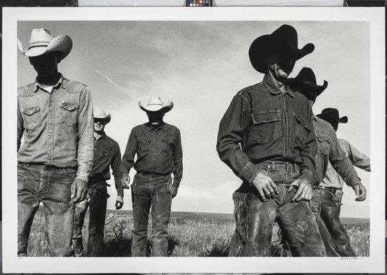 A black-and-white photograph of six cowboys walking in a field toward the viewer with their faces in shadow from their cowboy hats.