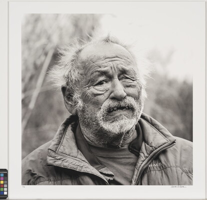 A black-and-white portrait photograph of a older light-skinned man, balding and with overgrown facial hair, wearing a quilted jacket standing outdoors.