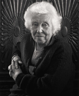 A black-and-white portrait photograph of an elderly White woman with fluffy white hair, posed in front of a carved wooden wall.