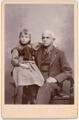 A black-and-white studio photograph of a seated White man with white hair and mustache wearing a three-piece suit and a young White girl in a sailor-style dress on his lap.