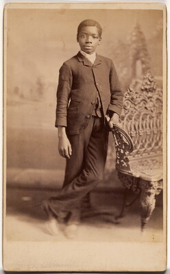 A sepia-toned studio portrait photograph of a Black boy wearing a suit, leaning against a wrought iron bench.