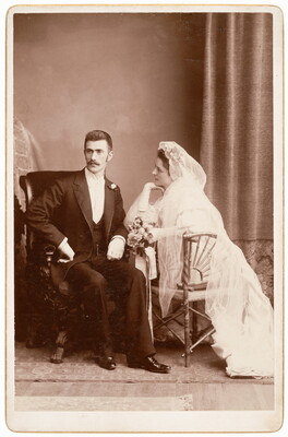 A black-and-white studio photograph of a White man wearing a tuxedo seated next to a White woman wearing a wedding dress with veil and holding flowers.