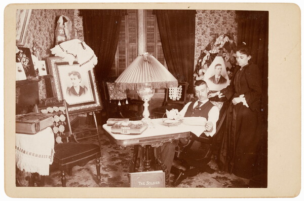 A black-and-white photograph of a parlor with a large table in the middle, a large lamp on top; a White man reads at the table as a White woman stands beside him looking over his shoulder.