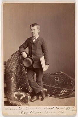 A sepia-toned photograph of a White boy in a suit, standing on a draped carpet, holding a notebook under one arm and a felt hat in his hand.