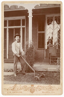 A black-and-white photograph of a White man with a mustache wearing bowler hat, bow tie, vest, and work gloves pushing a reel mower in front of a home.