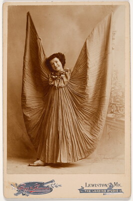 A sepia-toned studio photograph of a White woman wearing a long crinkled dress holding her arms up so the dress billows like wings.