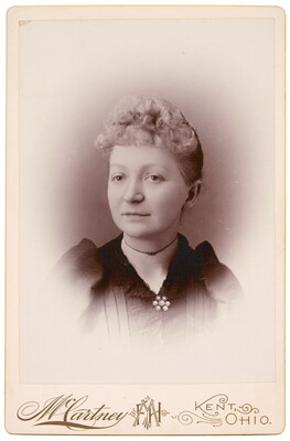 A sepia-toned studio portrait photograph of a White woman with curly, light colored hair in an updo, wearing a dark top closed with a brooch at the neck.