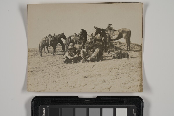 A sepia-toned photograph of a group of men sitting outside on the ground playing cards as their saddled horses stand behind them.