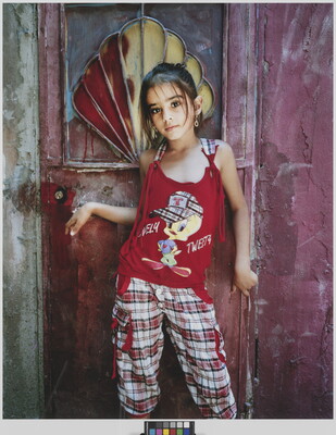 A color portrait photograph of a young medium-skinned girl wearing a red sleeveless shirt and red plaid pants standing in front of a door.