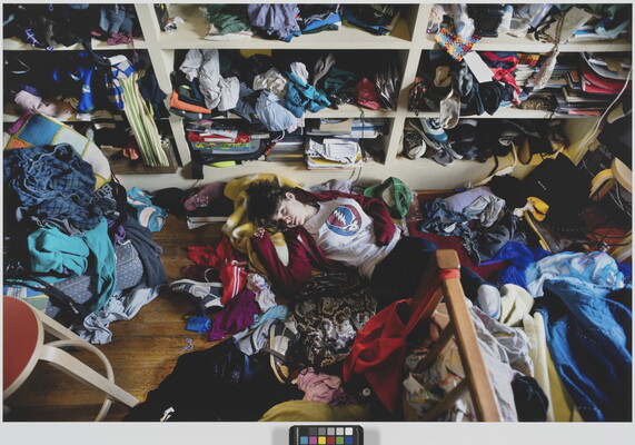 A color photograph of a light-skinned teenage girl lying on the floor of a messy room surrounded by unfolded clothing.
