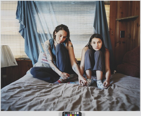 A color photograph of two light-skinned women holding hands and sitting on a bed in front of a fabric-covered window.