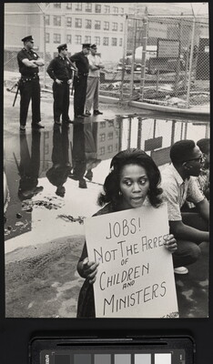 A black-and-white photograph of a Black woman holding a protest sign as White police officers stand in the background.