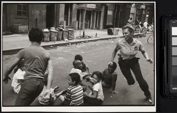 A black-and-white photograph of a smiling, White police woman playing a chase game with children of different races in a trash strewn street.
