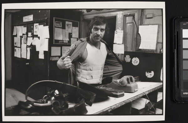 A black-and-white photograph of a White man standing behind a cluttered counter holding open his shirt to show a bullet-proof vest.