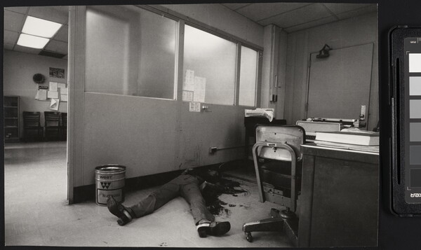 A black-and-white photograph of an office with a bloody body on the floor, feet toward the camera, the top part of the body obscured by dark stains on it, the floor, and the wall.