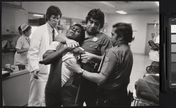 A black-and-white photograph of a Black man being restrained and choked by two police officers with medium to dark skin in a hospital setting.