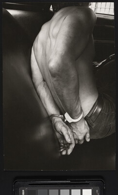 A black-and-white photograph of the bare back and handcuffed hands of a White man in the back seat of a police car.