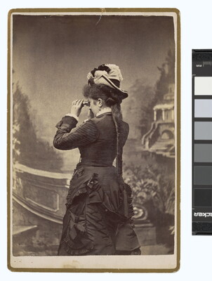 A sepia-toned studio photograph of a light-skinned woman, viewed from the back, wearing a dark corseted gown and peering through binoculars toward a painted landscape.