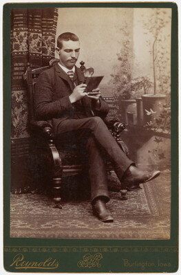 A black-and-white studio photograph of a seated White man using a magnifying glass to look at something he is holding.