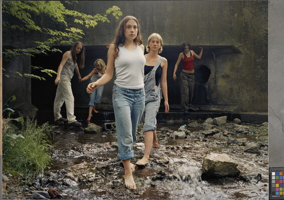 A color photograph of White teenagers, casually dressed and several barefoot, walking through a stream that emerges from a culvert behind them.