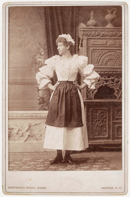 A black-and-white studio photograph of a young White person, hands on hips, with curly hair wearing a ruffled hat, blouse with puffy sleeves, and an apron over a dress.