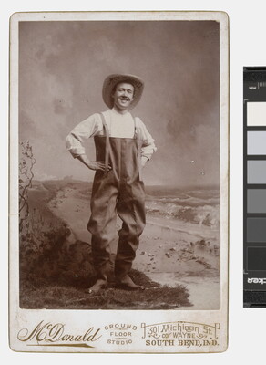 A sepia-toned studio photograph of a smiling White man wearing waders and a large straw hat, posed with hands on hips.